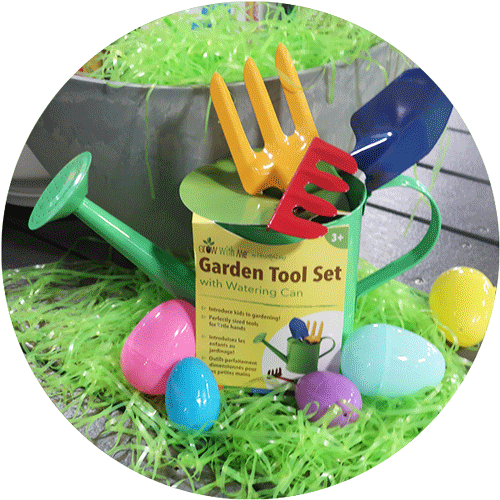 A green kids watering can with red, yellow and blue garden tools inside, surrounded by green easter grass and colorful easter eggs.